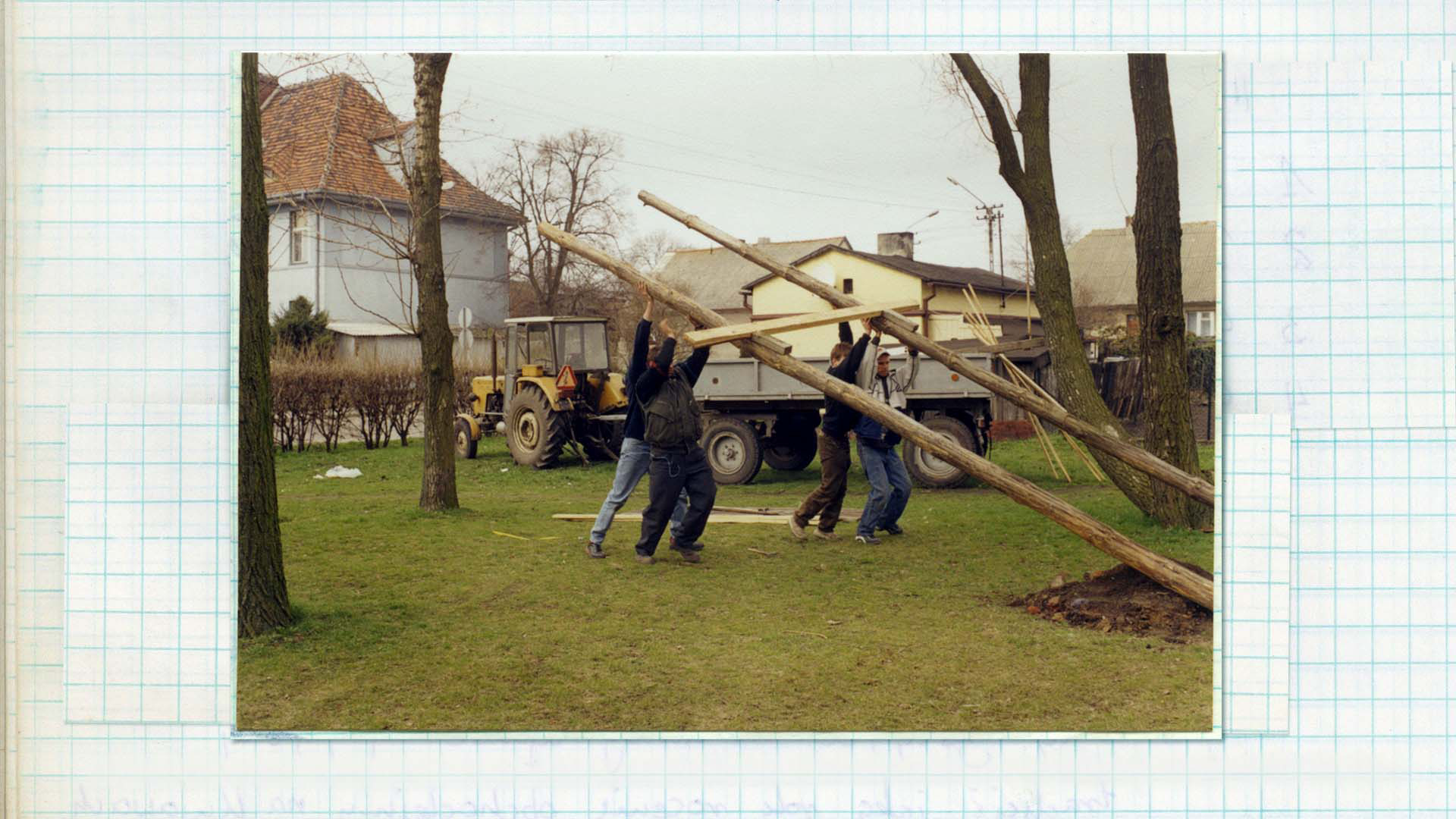 Fotografia w kolorze. Czwórka mężczyzn podczas ustawiania drewnianej konstrukcji, o dużych wymiarach. W tle traktor z przyczepą i budynki mieszkalne.