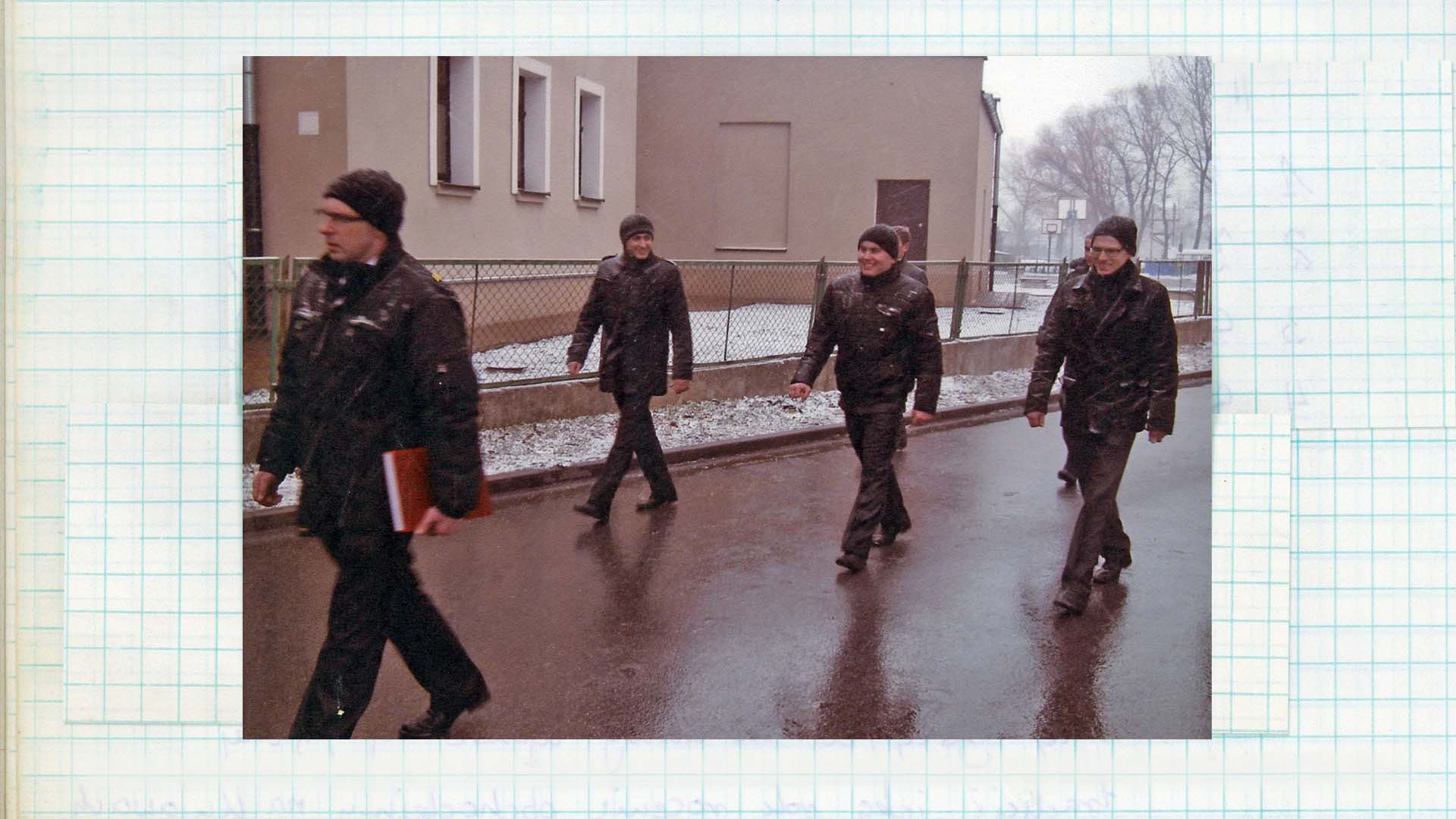 Zdjęcie w kolorze. Grupa mężczyzn przechodzi ulicami miejscowości. Mężczyźni uśmiechają się do siebie, a ich ubrania pokrywa cienka warstwa śniegu. W tle widoczne budynki i trawnik pokryty śniegiem.