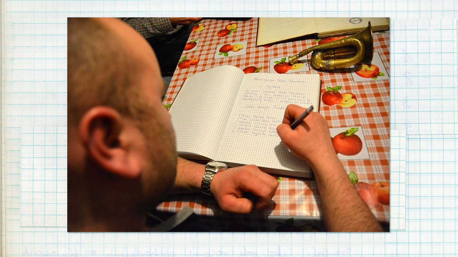 Układanie przywołówek , 2016, fot. Marek Jasik/ ino.online Zdjęcie kolorowe. Kadr przez ramię mężczyzny zapisującego notatki w papierowym dzienniku. Na stole leżą także; inny notes i złota trąbka.